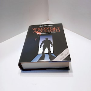 May include: A black book with the title "Wrongly Accused: The Maureen Prescott Murder" by Gale Weathers. The cover features a silhouette of a person holding a gun in front of a doorway. The book has a 25th Anniversary sticker on the bottom right corner.
