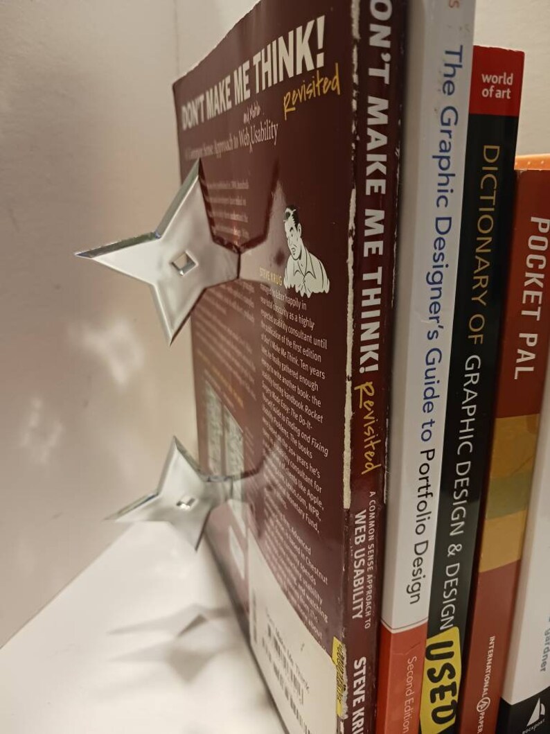 May include: A stack of books with a silver bookend shaped like a star. The books are titled "Don't Make Me Think! Revisited", "The Graphic Designer's Guide to Portfolio Design", "Dictionary of Graphic Design & Design", and "Pocket Pal".