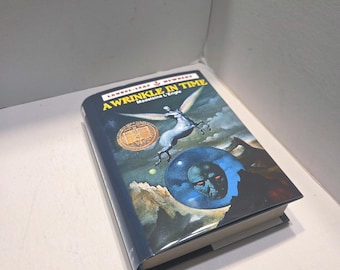May include: A hardcover book titled "A Wrinkle in Time" by Madeleine L'Engle. The cover features a blue and black illustration of a winged horse, a planet, and a shadowy figure. The book is on a white surface.