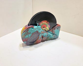 May include: A portable record player featuring a multi-colored paint splatter finish in shades of teal, red, and yellow. The device includes a circular speaker grille, a tone arm, and a black vinyl record with a colorful center label visible.