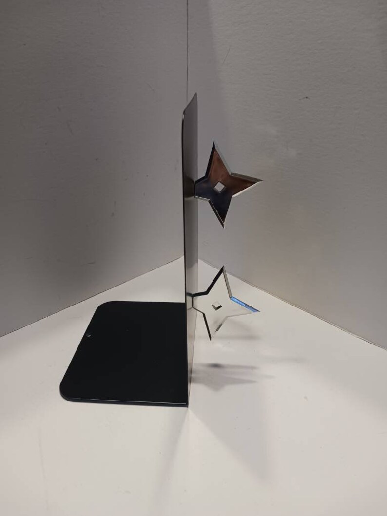 May include: A black metal bookend with two silver throwing stars attached to the side. The bookend is standing on a white surface.
