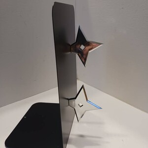 May include: A black metal bookend with two silver throwing stars attached to the front. The bookend is standing on a white surface.
