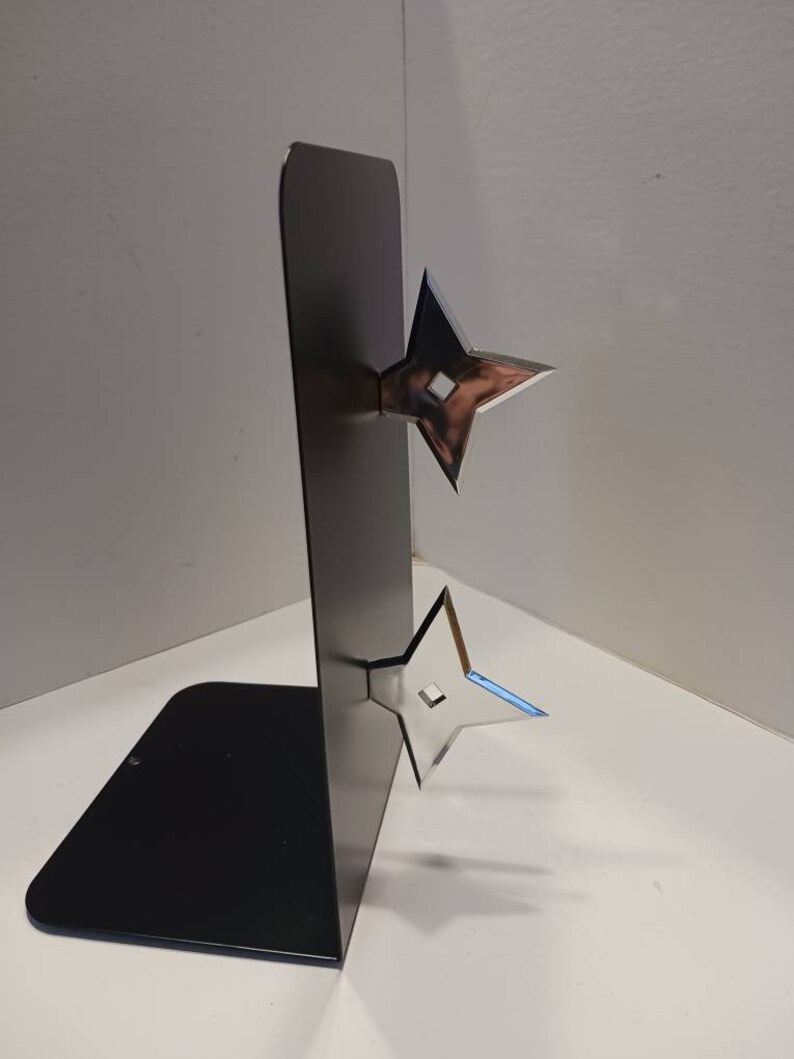 May include: A black metal bookend with two silver throwing stars attached to the front. The bookend is standing on a white surface.