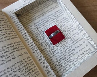 May include: A silver ring rests in a red velvet box, nestled inside a hollowed-out book. The book's pages are visible, with text and a rectangular cut-out. The ring is the focal point.