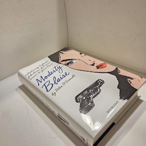 May include: A hardcover book titled "Modesty Blaise" by Peter O'Donnell. The cover features a stylized illustration of a woman's face, a handgun, and the publisher's name, Doubleday. The book is white with blue and black text.