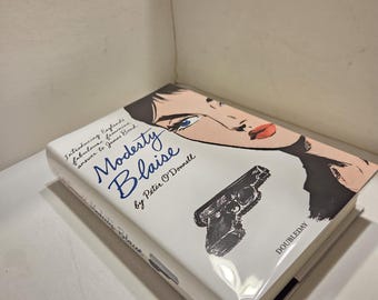 May include: A hardcover book titled "Modesty Blaise" by Peter O'Donnell. The cover features a stylized illustration of a woman's face, a handgun, and the publisher's name, Doubleday. The book is white with blue and black text.
