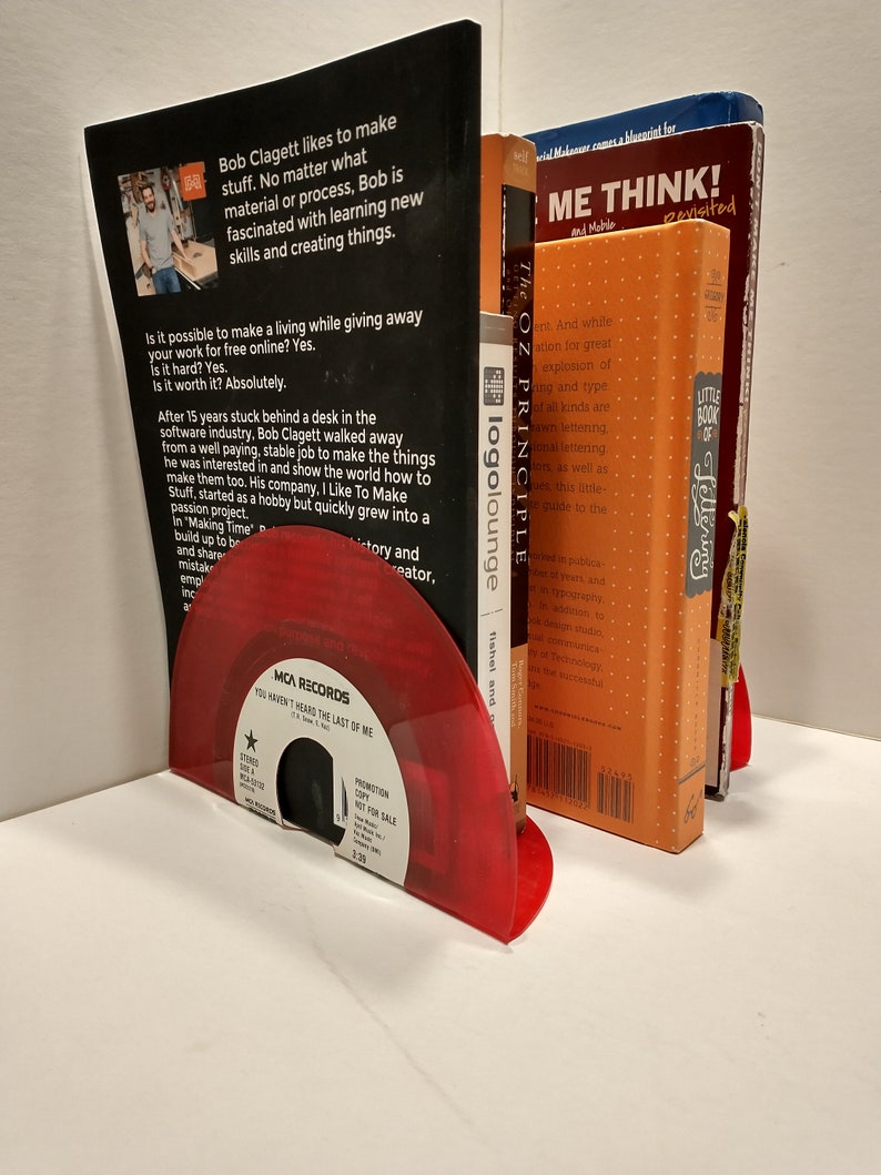 Small, shiny, round 45 size, vinyl records are bent on the bottom to make bookends that holds up some bright, colorful books. The color of the album is red.