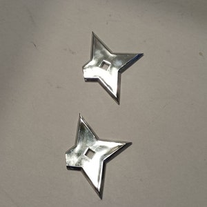 May include: Two silver throwing stars, also known as shurikens, are shown on a white surface. The stars have four points and a small hole in the center.