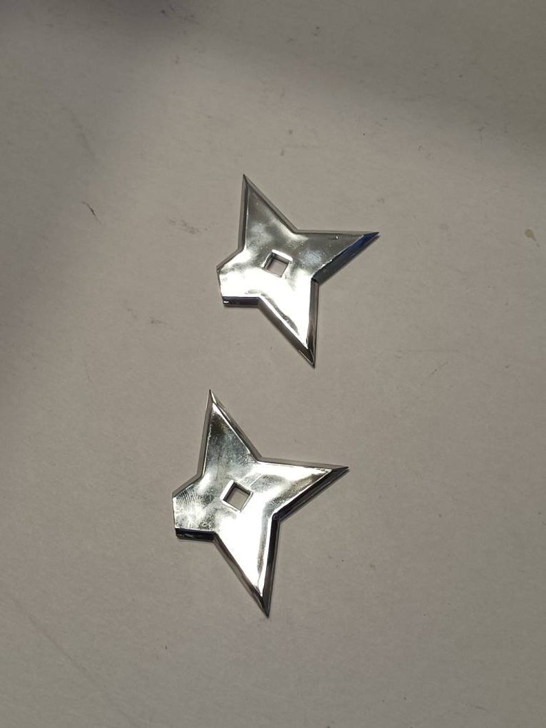 May include: Two silver throwing stars, also known as shurikens, are shown on a white surface. The stars have four points and a small hole in the center.
