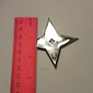May include: A silver-colored throwing star, also known as a shuriken, is shown against a white background. A red ruler is visible in the image, with the throwing star measuring approximately 2 inches in diameter.