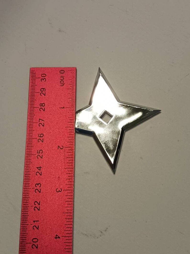 May include: A silver-colored throwing star, also known as a shuriken, is shown against a white background. A red ruler is visible in the image, with the throwing star measuring approximately 2 inches in diameter.