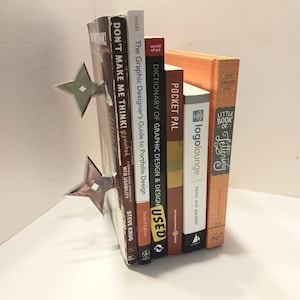 May include: A set of six books with a silver bookend shaped like a throwing star. The books are stacked vertically and include titles like "Don't Make Me Think!" and "The Graphic Designer's Guide to Portfolio Design".