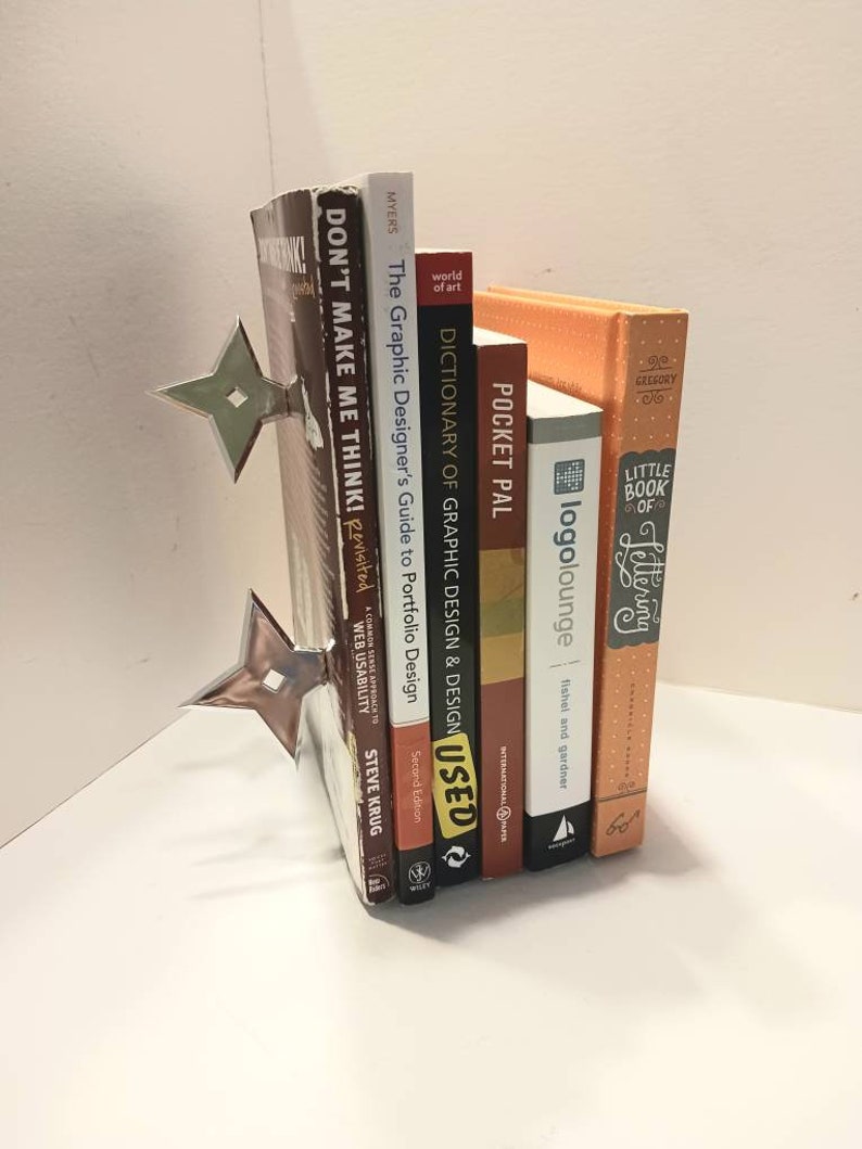 May include: A set of six books with a silver bookend shaped like a throwing star. The books are stacked vertically and include titles like "Don't Make Me Think!" and "The Graphic Designer's Guide to Portfolio Design".