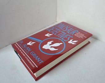 May include: A hardcover book titled "Dinosaur Detectives" with a red cover. The cover features a blue circle with a dinosaur footprint and three smaller footprints. The author's name, Dr. Alan Grant, is printed in white.