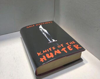 May include: A hardcover book titled "Knife of the Hunter" by Gale Weathers. The book has a black cover with orange lettering and a graphic of a knife. The book's pages are a light tan color.