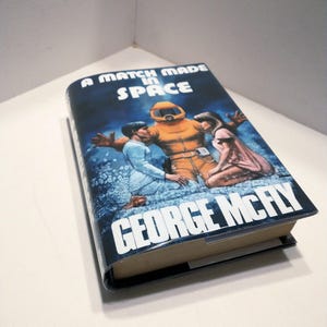 May include: A book cover with a blue background and the title "A Match Made in Space" in white text. The cover features an illustration of three people in space suits, with one person in a yellow suit and two others in blue suits.