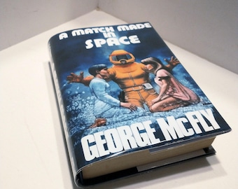May include: A book cover with a blue background and the title "A Match Made in Space" in white text. The cover features an illustration of three people in space suits, with one person in a yellow suit and two others in blue suits.