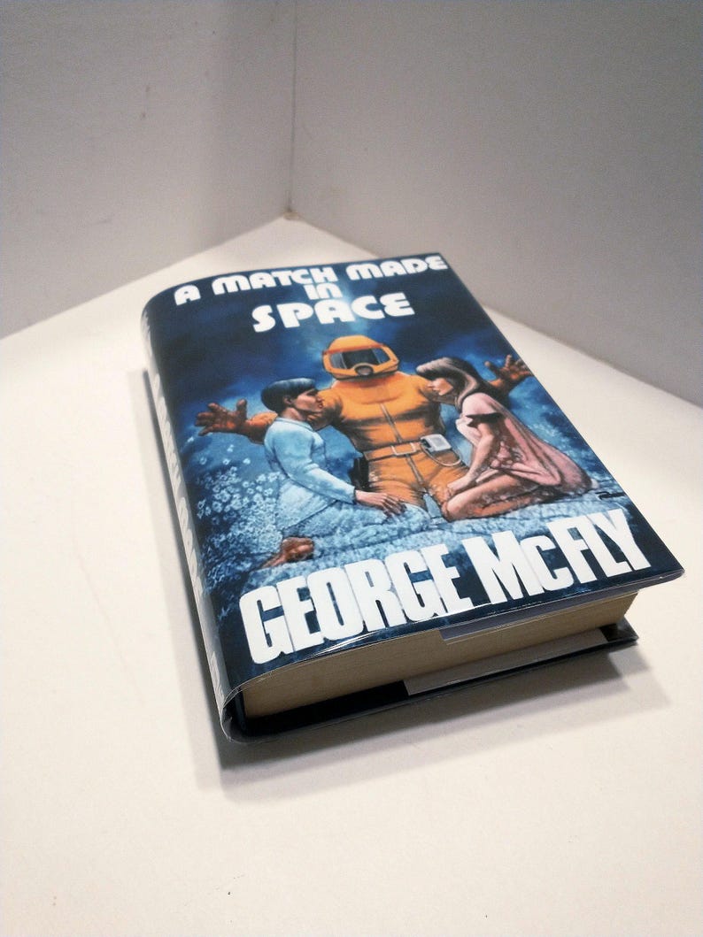May include: A book cover with a blue background and the title "A Match Made in Space" in white text. The cover features an illustration of three people in space suits, with one person in a yellow suit and two others in blue suits.