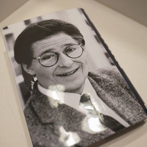 May include: Black and white photograph of a person wearing glasses and a suit jacket. The person is smiling and looking at the camera.