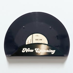 May include: A black vinyl record displayed in a semi-circular holder. The record label reads "Baby Jane". The holder has the words "Now Spinning" in white script. The background is plain white.