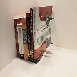 May include: A stack of books on a white shelf. The top book is red and has the title "The Graphic Designer's Portfolio Design" on the cover. The other books are various colors and have titles like "Don't Make Me Think!" and "Dictionary of Graphic Design & Design".