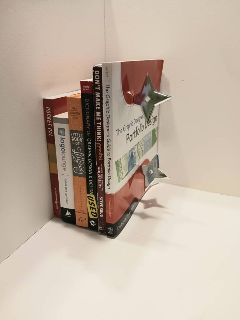 May include: A stack of books on a white shelf. The top book is red and has the title "The Graphic Designer's Portfolio Design" on the cover. The other books are various colors and have titles like "Don't Make Me Think!" and "Dictionary of Graphic Design & Design".