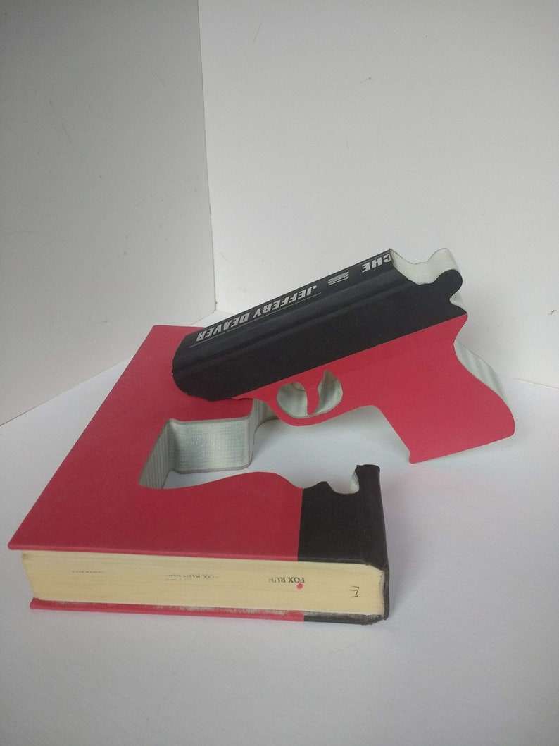 May include: A red book with a black spine and the title "The Jeffery Deaver" is cut out to form the shape of a gun. The gun is black and red.