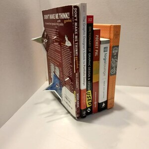 May include: A stack of six books about graphic design, web design, and lettering. The books are arranged vertically with the tallest book on the left and the shortest book on the right. The titles of the books are visible: "Don't Make Me Think!" "Don't Make Me Think!" "The Graphic Designer's Guide to Portfolio Design" "Dictionary of Graphic Design & Design" "Pocket Pal" "Little Book of Lettering" and "logolounge".