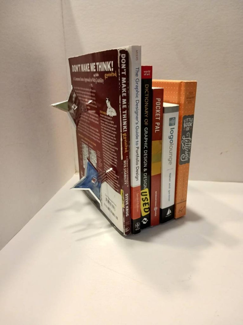 May include: A stack of six books about graphic design, web design, and lettering. The books are arranged vertically with the tallest book on the left and the shortest book on the right. The titles of the books are visible: "Don't Make Me Think!" "Don't Make Me Think!" "The Graphic Designer's Guide to Portfolio Design" "Dictionary of Graphic Design & Design" "Pocket Pal" "Little Book of Lettering" and "logolounge".