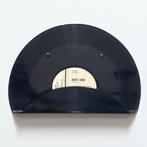 May include: A black vinyl record shelf with a curved design. The shelf holds a 45 RPM record with the label "Baby Jane" visible. The shelf is mounted on a white wall, creating a minimalist aesthetic.