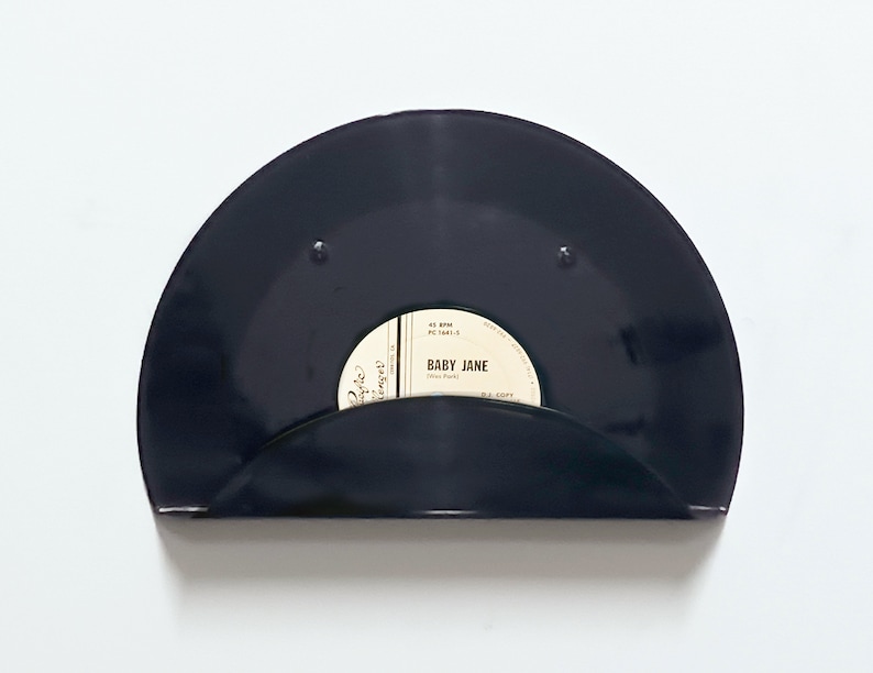 May include: A black vinyl record shelf with a curved design. The shelf holds a 45 RPM record with the label "Baby Jane" visible. The shelf is mounted on a white wall, creating a minimalist aesthetic.
