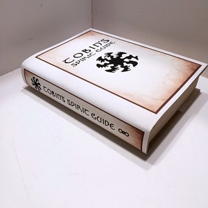 May include: A white book with a brown spine and a black and white illustration of a symbol on the cover. The title of the book is "Cobin's Spiric Guide".