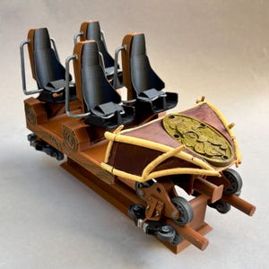 Può includere: Modello dettagliato di un vagone di montagne russe con quattro sedili neri e barre di sicurezza argentate. Il vagone è marrone con dettagli dorati e uno scudo decorativo. Le ruote sono grigie e il design è intricato.