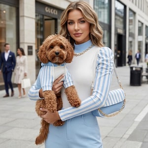 Może przedstawiać: Kobieta trzyma małego brązowego psa w niebiesko-białej bluzie z kapturem w paski. Kobieta ma na sobie białą górę z niebiesko-białymi paskami na rękawach, jasnoniebieskie spodnie i pasującą torebkę. Złoty naszyjnik z łańcuszka i sklep Gucci są widoczne w tle.