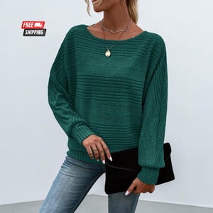 May include: A dark green long-sleeve sweater featuring a horizontal ribbed texture on the torso and vertical ribbed detailing on the sleeves. The sweater has a wide, rounded neckline and is paired with blue denim jeans and a black clutch bag.