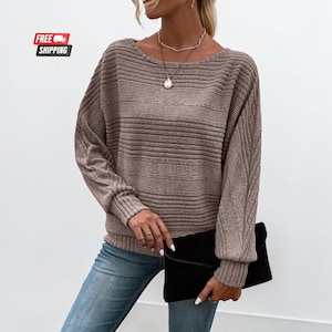 May include: A long-sleeved, taupe-colored sweater featuring a horizontal ribbed texture on the torso and a diagonal ribbed pattern on the sleeves. The garment has a wide, rounded neckline and is paired with blue denim jeans and a black clutch.
