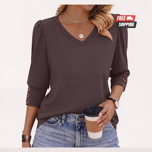 May include: A dark brown V-neck top with three-quarter length sleeves featuring three buttons on each cuff. The top is paired with blue denim jeans. A gold layered necklace with a circular pendant and a wristwatch are worn with the garment.