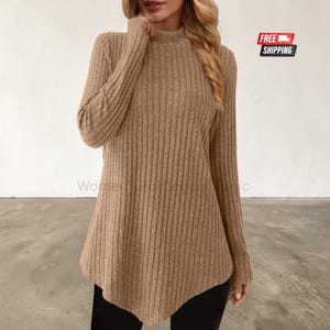 May include: A long-sleeved, tan-coloured jumper featuring a ribbed texture and a mock neck collar. The garment has a slightly flared, asymmetrical hemline that extends past the waist, providing a relaxed fit throughout the torso and arms.
