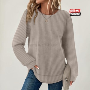 May include: A long-sleeved beige waffle knit tunic featuring a crew neckline and a curved hemline. The garment has a textured fabric surface and is paired with blue denim jeans. A delicate gold-toned necklace with a small pendant is worn.