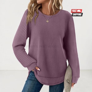 Peut inclure: Un pull à manches longues en maille gaufrée de couleur mauve avec une encolure ronde et un ourlet arrondi. Le vêtement présente un motif texturé à côtes verticales sur toute la surface et est associé à un jean bleu.