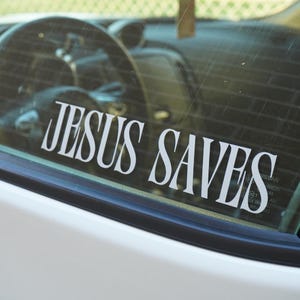 Könnte beinhalten: Weißer Aufkleber mit dem Schriftzug "JESUS SAVES" in einer stilisierten Schriftart, angebracht an der Innenseite einer Autoscheibe. Das Innere des Autos, einschließlich Lenkrad und Armaturenbrett, ist durch das Glas sichtbar.