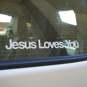 Puede incluir: Un coche blanco con una pegatina en la luneta trasera que dice "Jesus Loves You".
