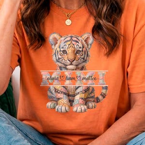 Può includere: T-shirt arancione con grafica di cucciolo di tigre, la parola "MOM" e nomi personalizzati. Il design del cucciolo di tigre è dettagliato e colorato. La maglietta è abbinata a jeans blu.