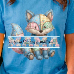 Può includere: T-shirt azzurra con un disegno di volpe patchwork. La volpe è realizzata con vari colori pastello. Sotto la volpe c'è la parola "NIMA" con i nomi "Olivia", "Liam" e "Matteo".