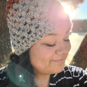 May include: A close-up of a person wearing a crocheted hat with a brown and grey ombre color scheme. The hat is made with a chunky yarn and has a textured stitch pattern. The person is wearing a striped shirt and has their eyes closed.