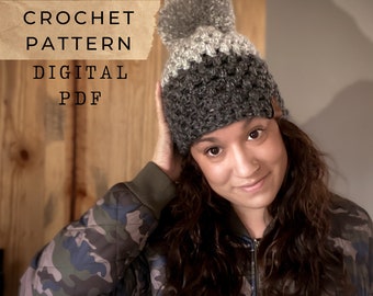 Puede incluir: Un patrón de gorro de crochet en formato PDF digital. El gorro es gris y blanco con un pompón en la parte superior. El patrón está disponible para descargar.