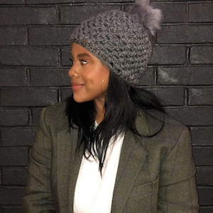 May include: A woman wearing a gray crocheted beanie with a faux fur pom pom. She is wearing a green blazer over a white shirt.
