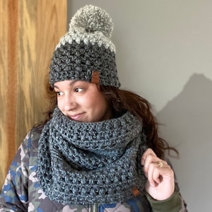 May include: A gray and white crocheted beanie with a pom pom on top. The beanie is worn by a person wearing a gray crocheted infinity scarf and a camouflage jacket.