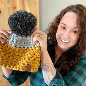 May include: A crocheted beanie with a gray and yellow color scheme and a black pom pom on top. The beanie has a brown leather tag with the word "Cozy" on it.