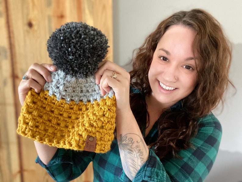 May include: A crocheted beanie with a gray and yellow color scheme and a black pom pom on top. The beanie has a brown leather tag with the word "Cozy" on it.